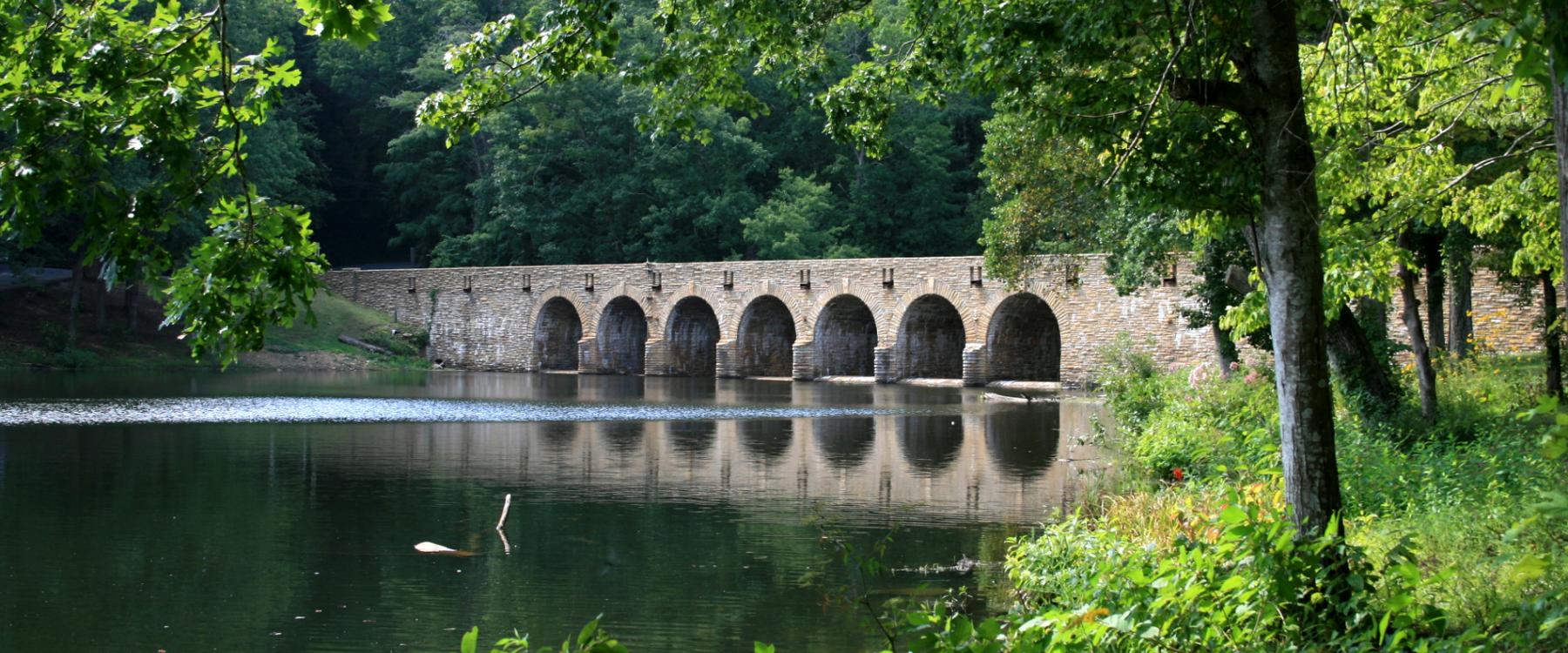 The same long arched stone bridge spans a calm lake, surrounded by dense green summer foliage.