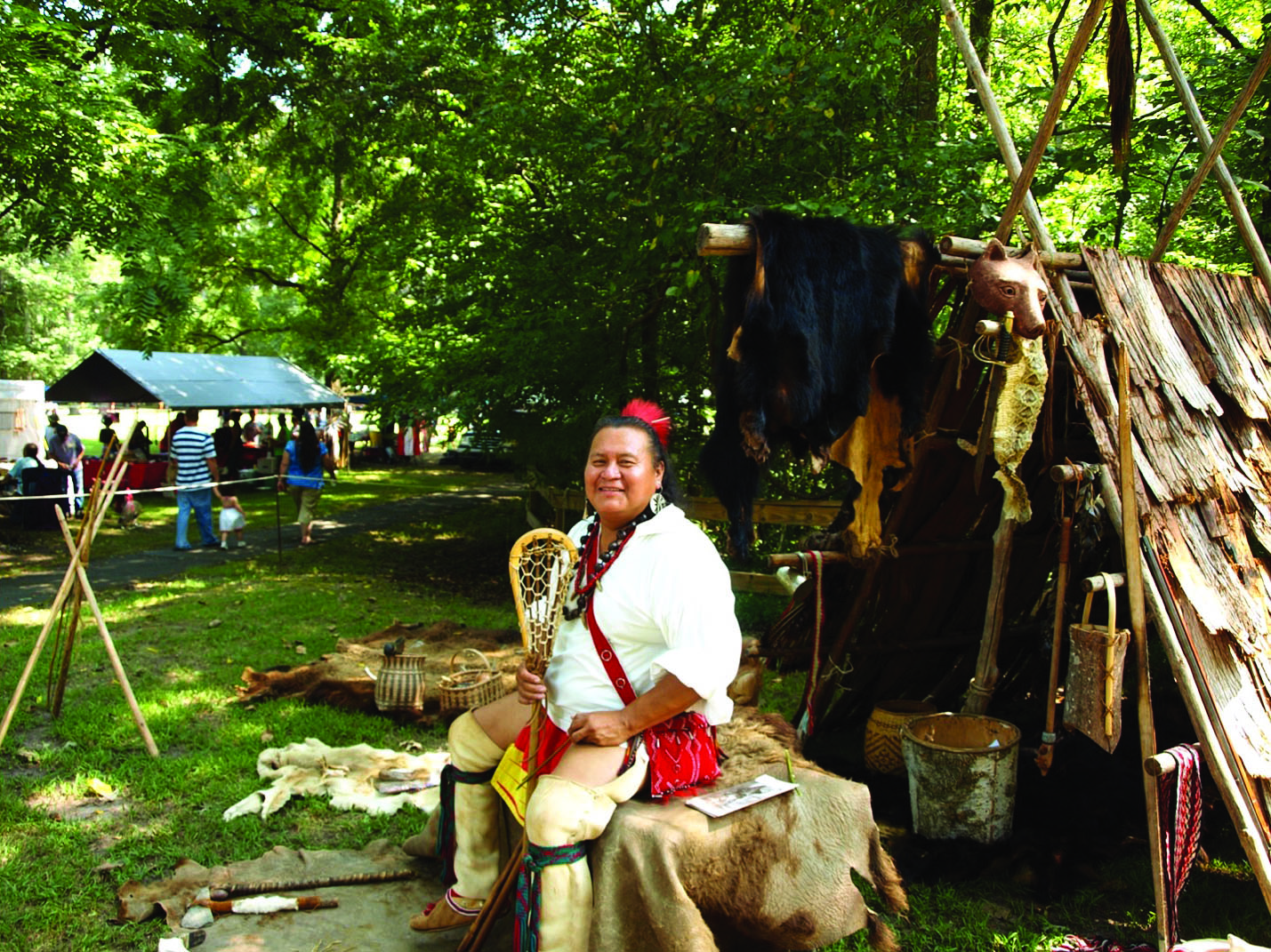 Person in traditional attire sits by rustic shelter with forest and small gathering nearby.