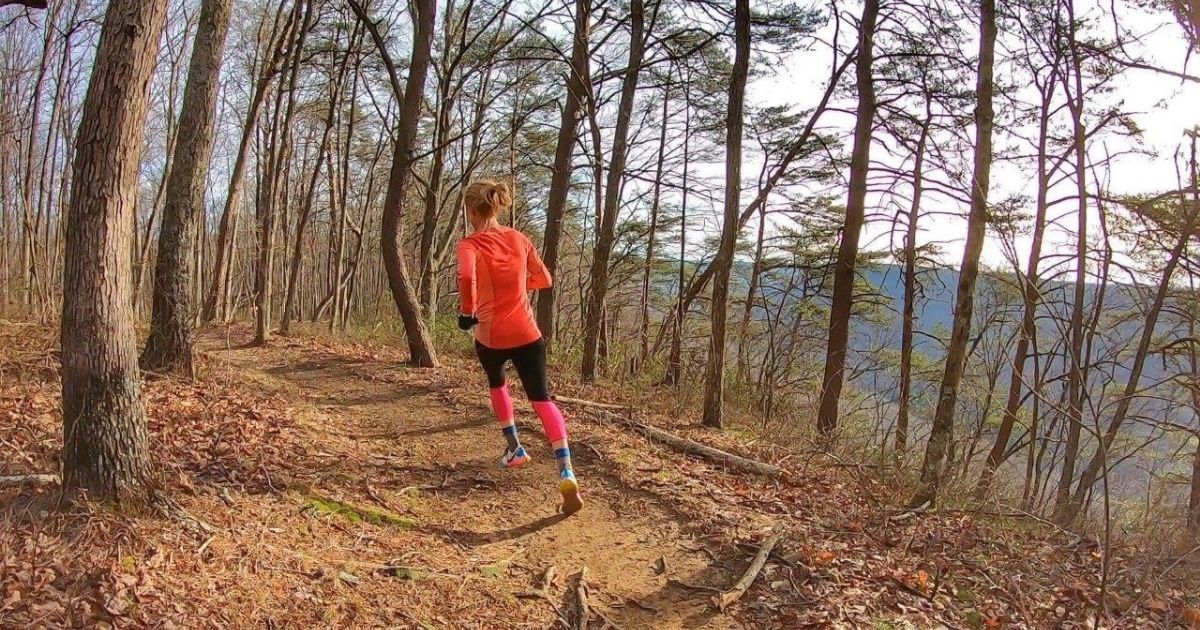 Trail runner in orange shirt and pink socks on a wooded path.