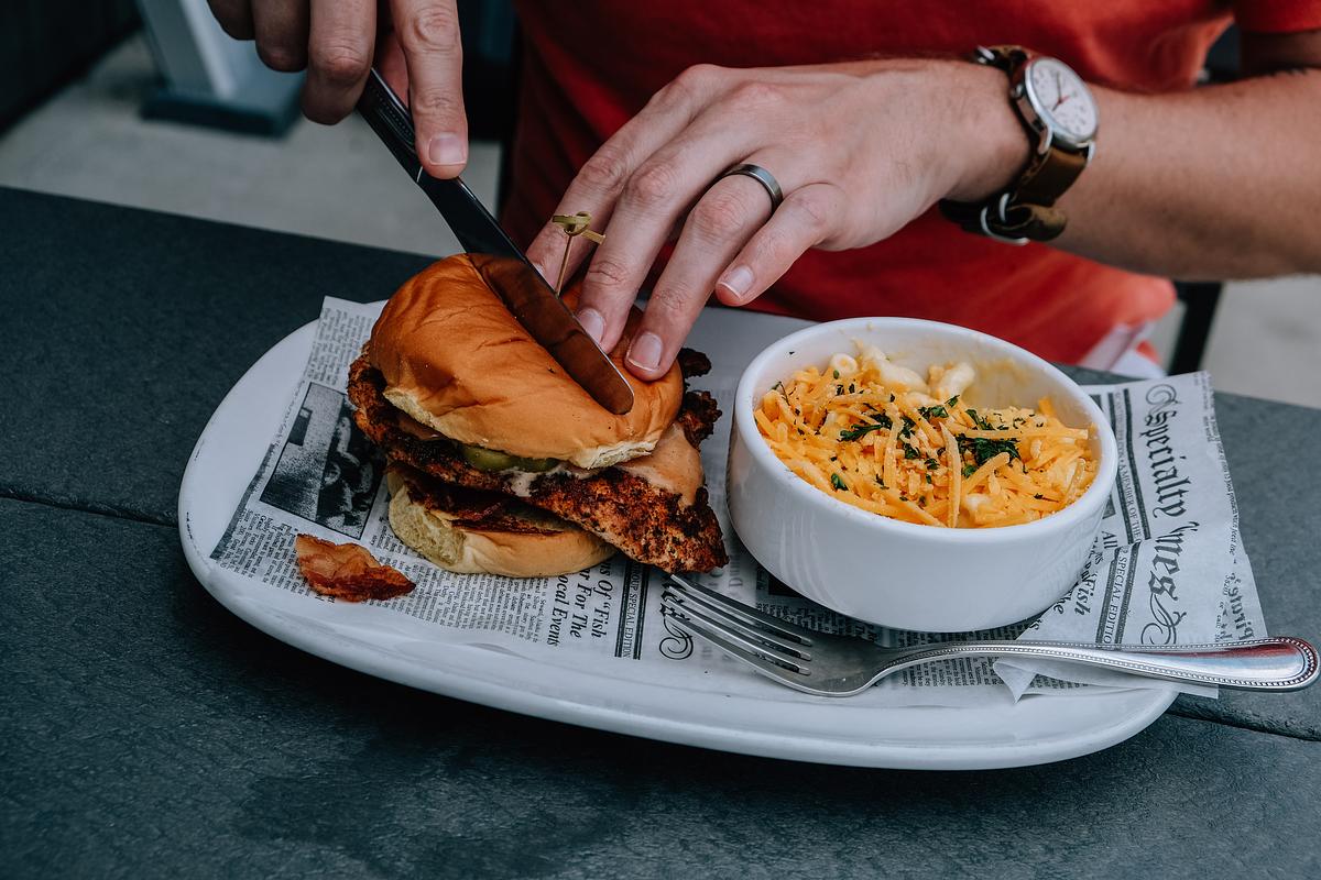 Person cutting a chicken sandwich, served with coleslaw on a newspaper-lined plate.