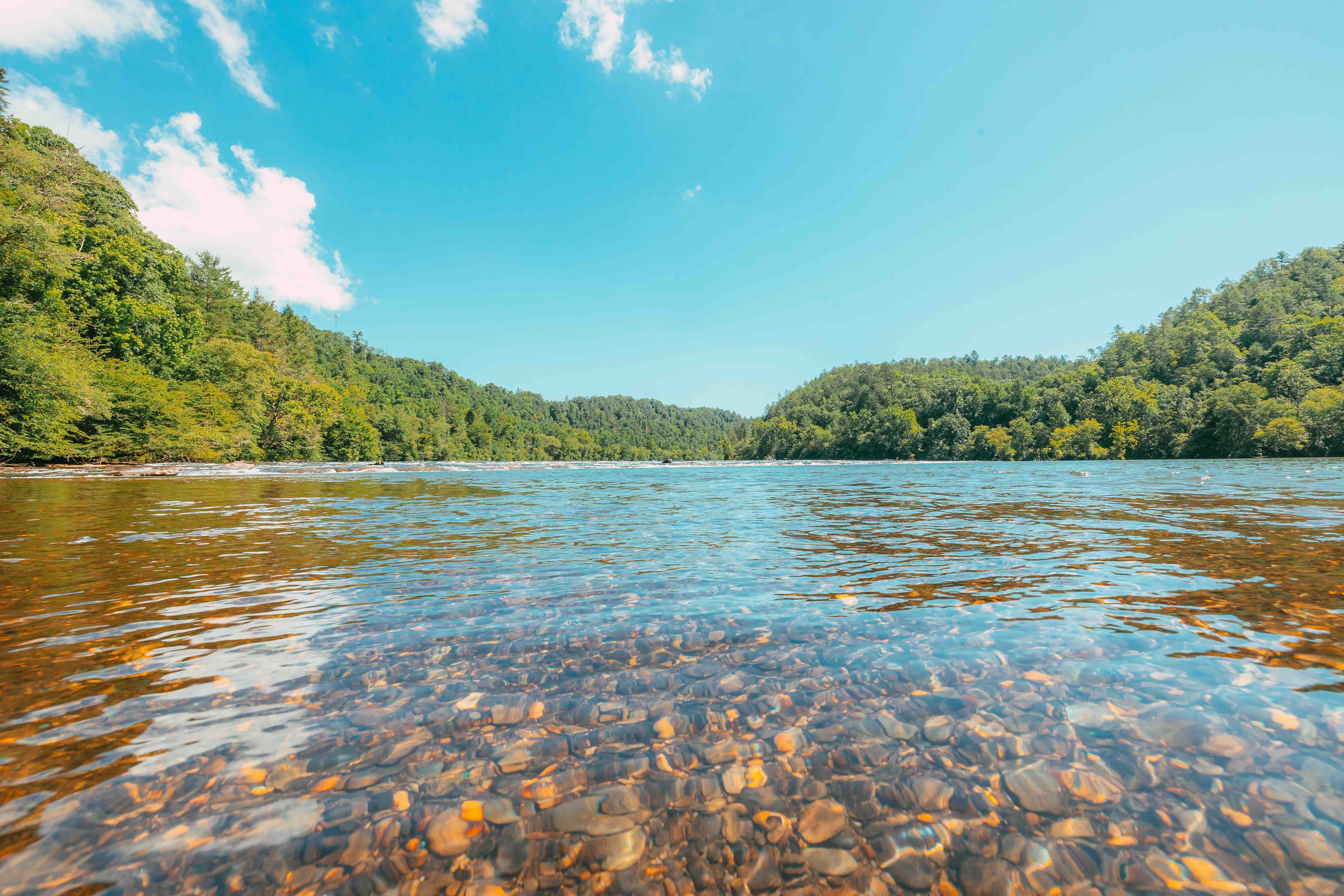 Clear river with rocky bed surrounded by lush green hills under a blue sky.