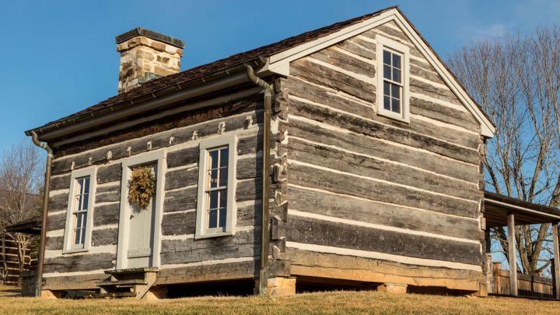 Rustic log cabin with chimney, set on a grassy hill.