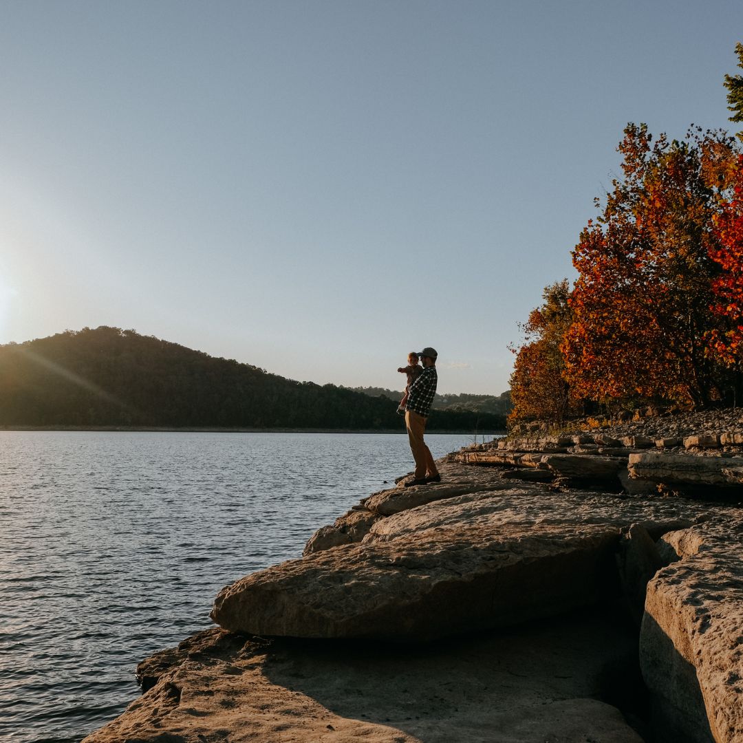 Person standing on a rocky shore, photographing sunset over a lake.