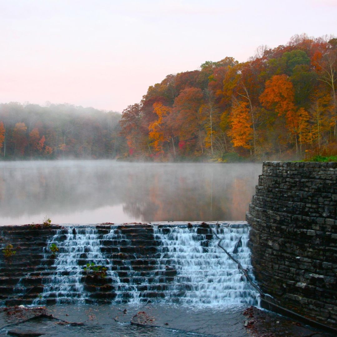 Waterfall over a stone dam with misty autumn trees in the background.