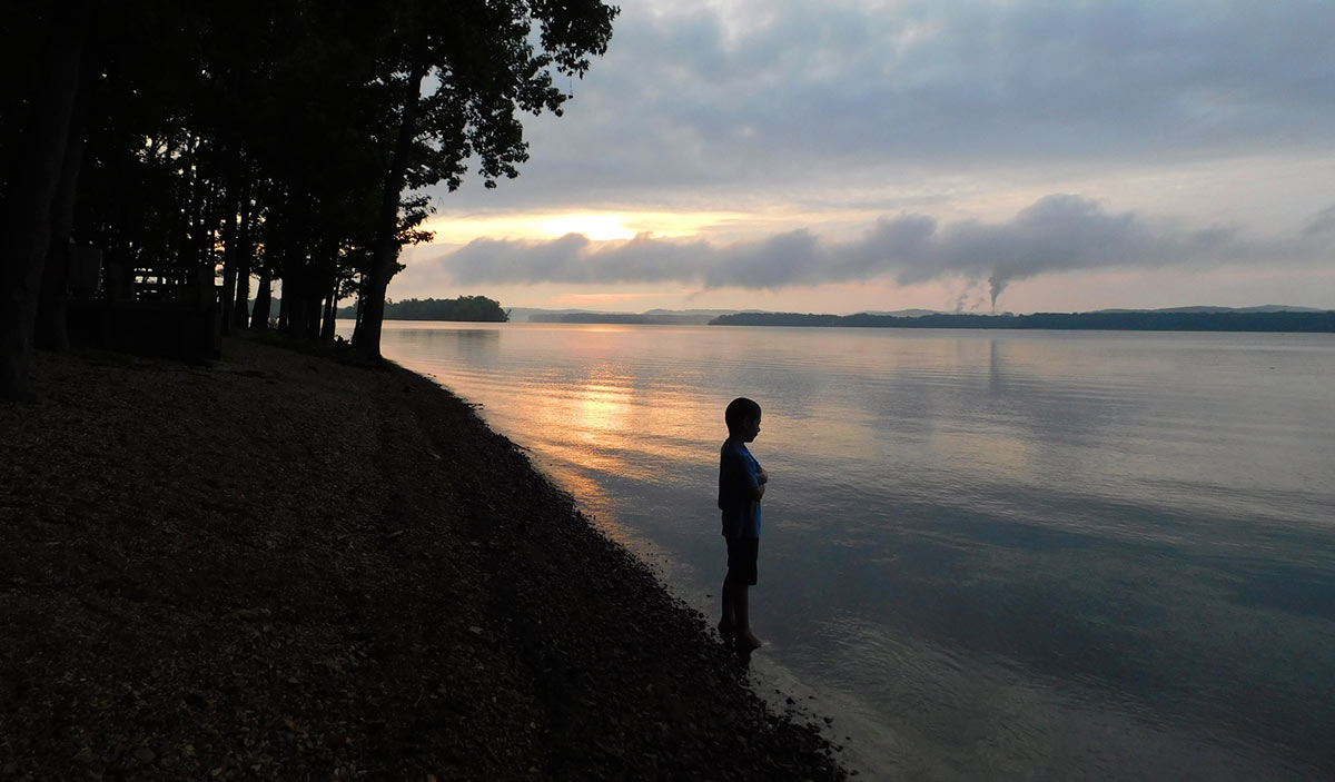 Silhouette of a person at lakeside during sunset with trees on the left.