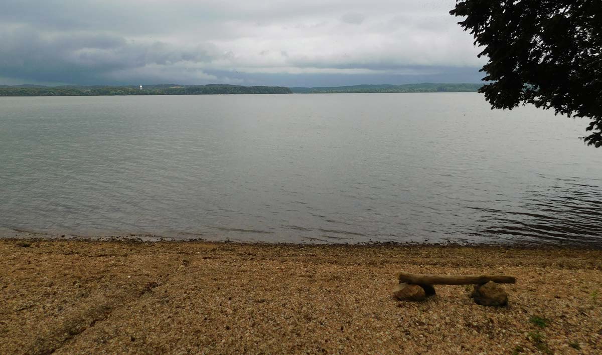 Pebble beach with a wooden bench, calm water, cloudy sky.
