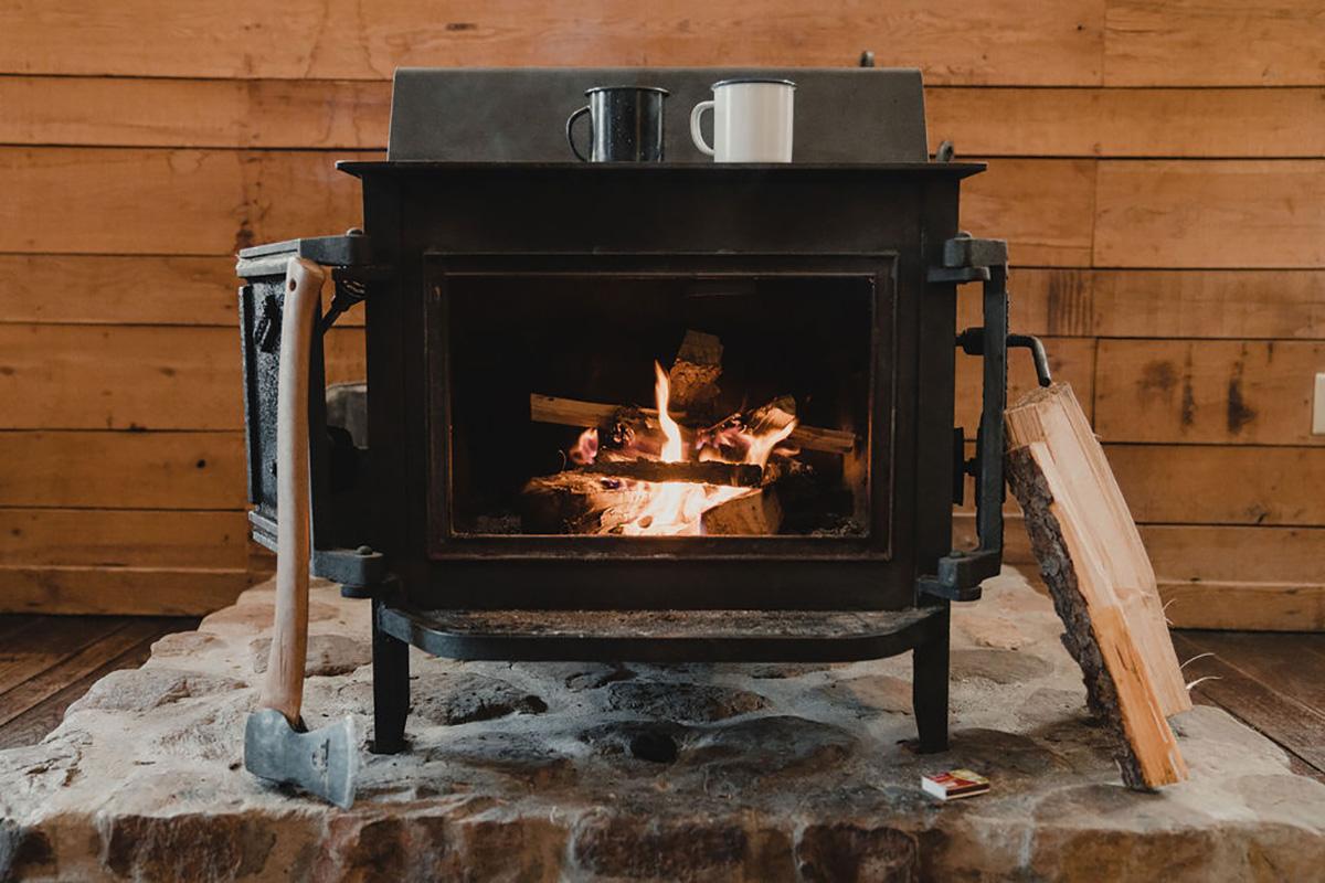 Wood-burning stove inside a cozy cabin, with a fire glowing behind the glass door. An axe leans against the stove, split logs sit nearby, and two mugs rest on top against a wooden wall backdrop.