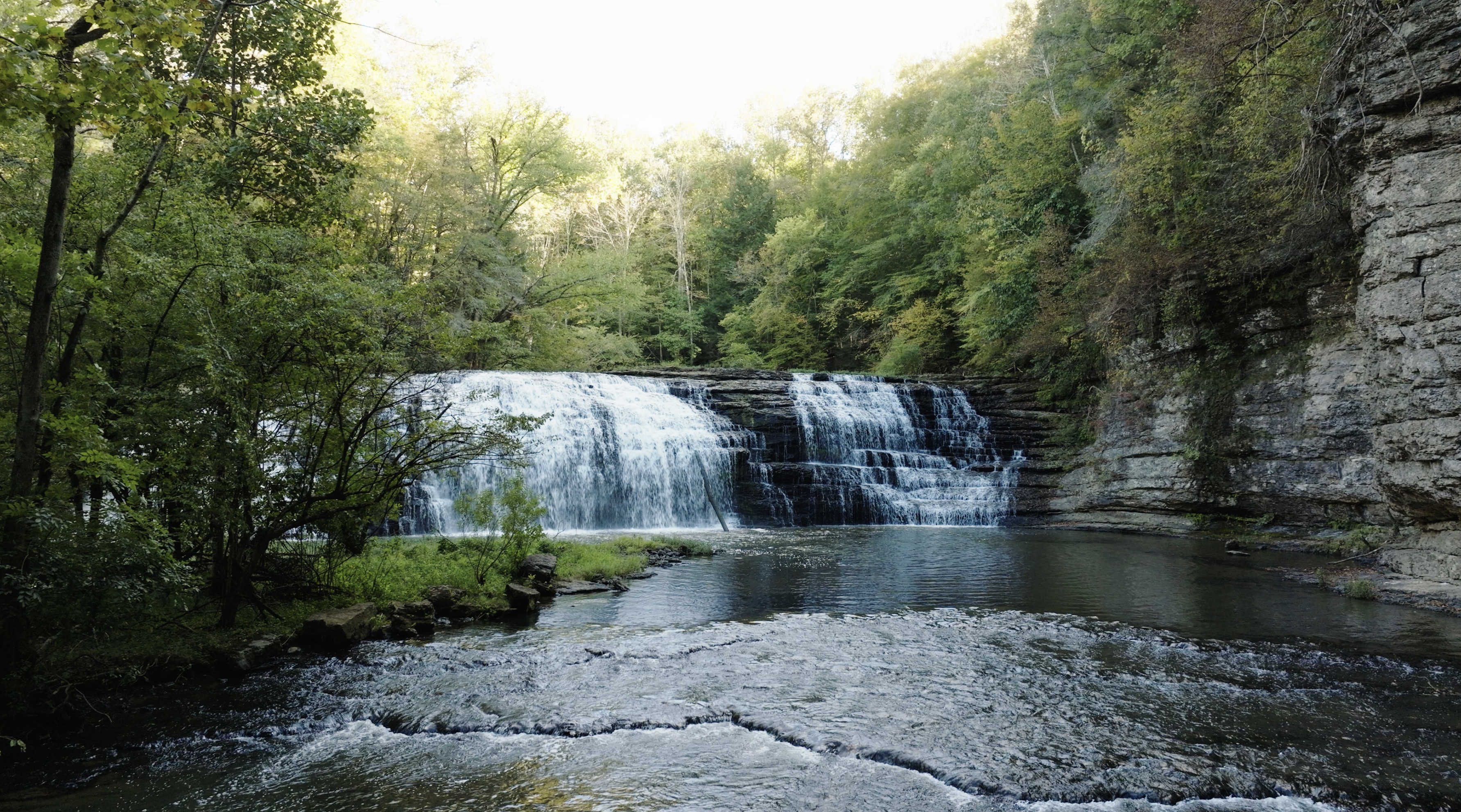 A cascading waterfall drops into a shallow pool surrounded by green trees