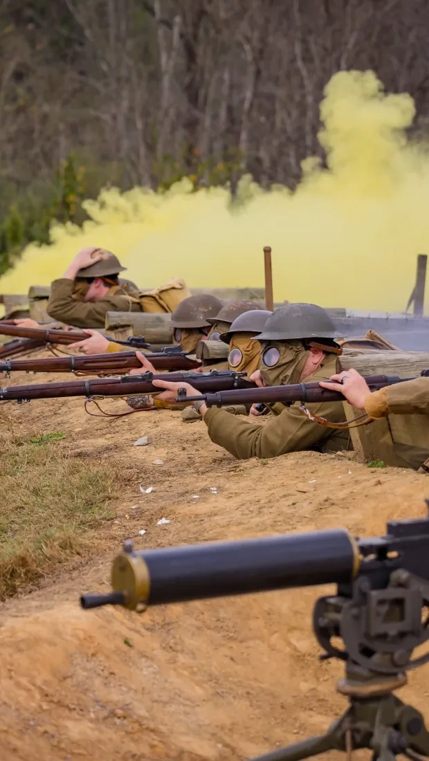 World War I reenactment scene with soldiers in helmets and gas masks aiming rifles from a trench, surrounded by yellow smoke, evoking tension.