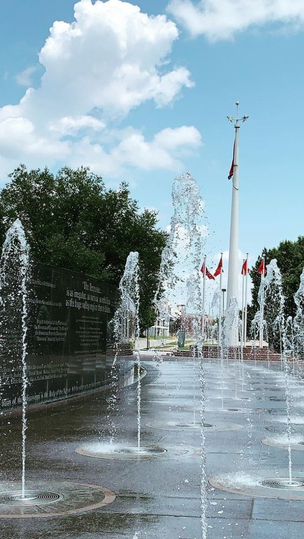 Fountains spray water upward in a plaza surrounded by green trees and a tall flagpole with flags. The sky is blue with fluffy clouds, creating a lively atmosphere.