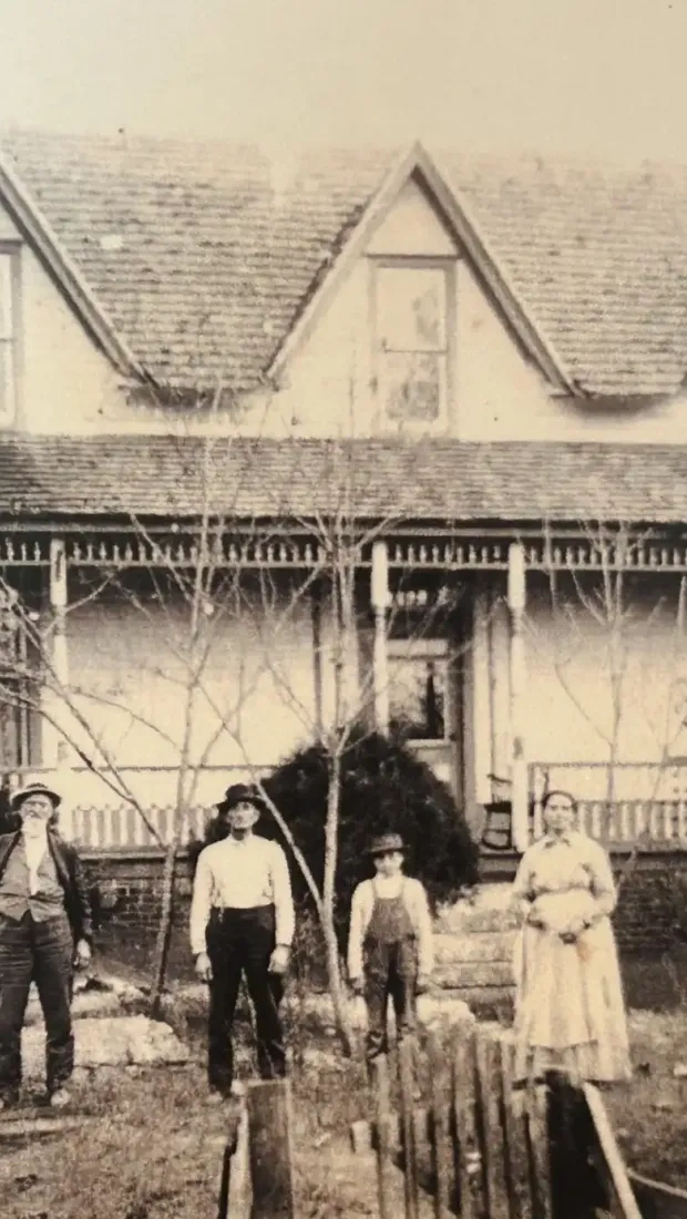 Four people stand in front of a vintage house with a porch and dormer windows.