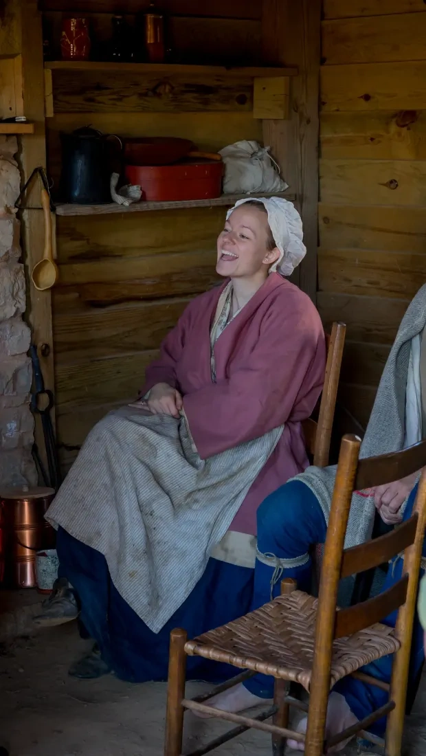 A group in historical attire sits in a rustic wooden cabin by a stone fireplace. They appear to be engaging in lively, cheerful conversation.