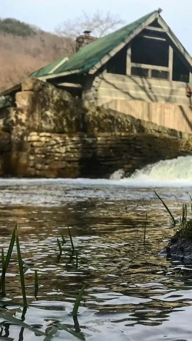 Old mill by a river with a gentle waterfall, surrounded by hills and grassy banks.