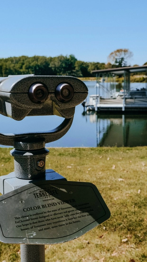 A coin-operated viewing machine stands on grass, overlooking a calm lake with a dock and boats. The scene is serene under a clear blue sky.