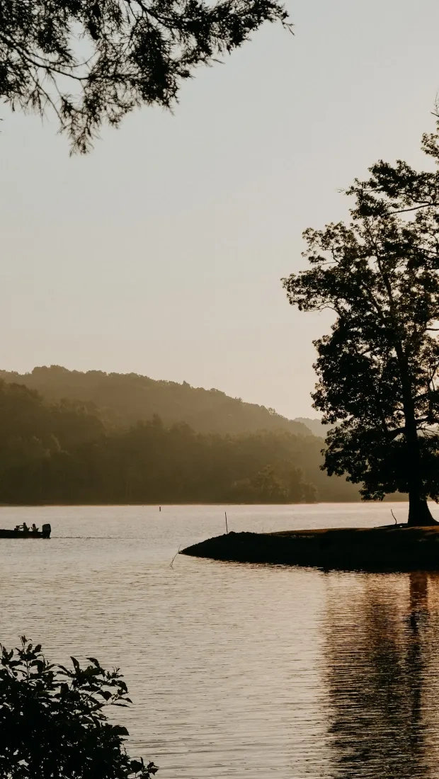 A small boat with passengers moves across a lake at sunset, with tree silhouettes and golden reflections on the water.