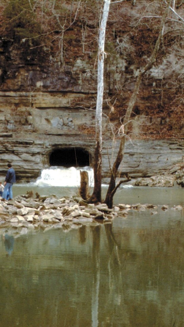 A serene scene of three people standing on a rocky riverbank near leafless trees, facing a cave opening with water flowing out, creating a peaceful atmosphere.