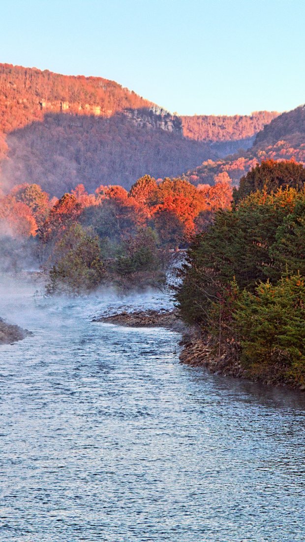 A serene river flows through a valley with vibrant autumn foliage on hills. Mist rises from the water, creating a peaceful and tranquil atmosphere.