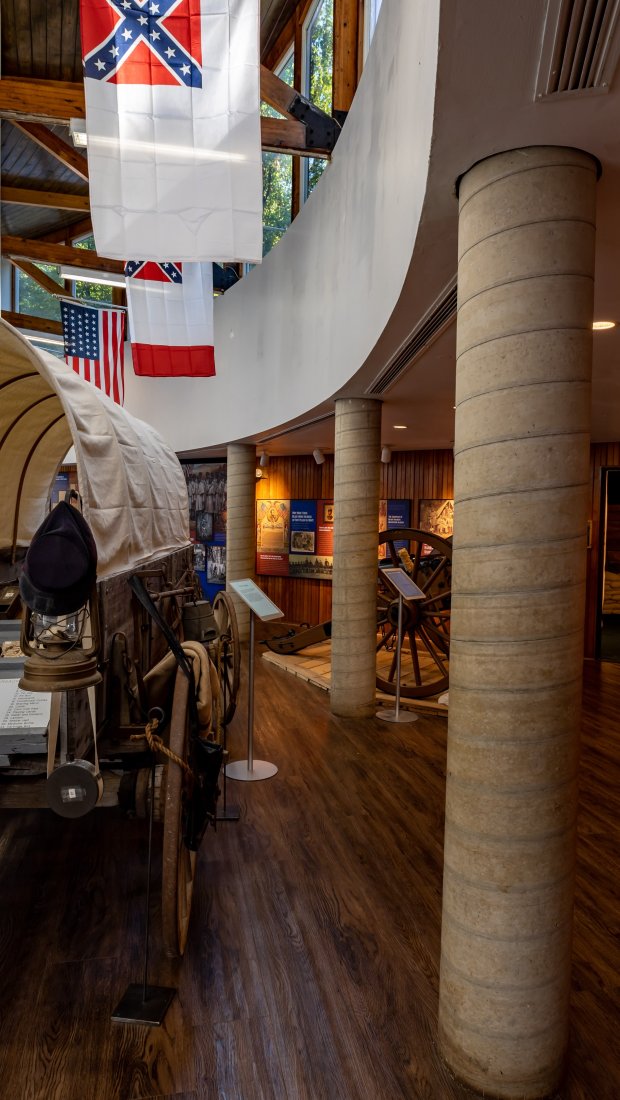 A historical exhibit shows a covered wagon with hats on its side. Flags hang overhead. Informational displays line the wooden walls, creating an educational atmosphere.