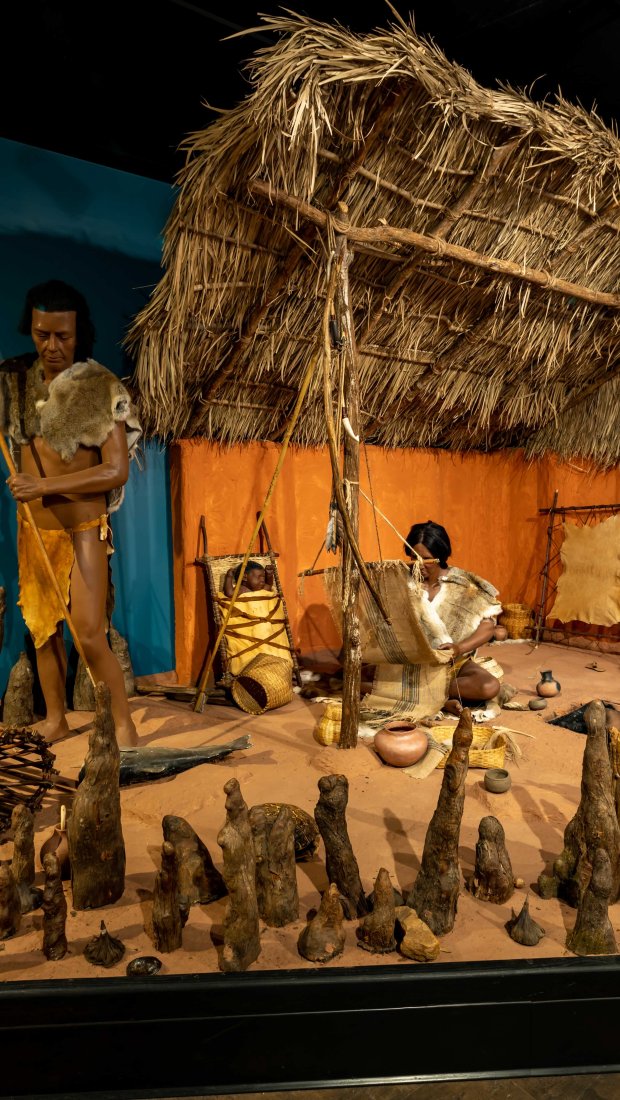  Museum diorama of early humans in a prehistoric setting. Two figures in fur clothing with tools, a thatched hut, clay pots, and surrounding stalagmites.