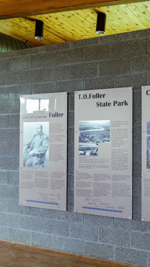 Interior of a modern building with gray brick walls featuring three informational panels about T.O. Fuller State Park, Fuller, and Chucalissa/Boxtown. The wooden ceiling and doors add warmth to the space.