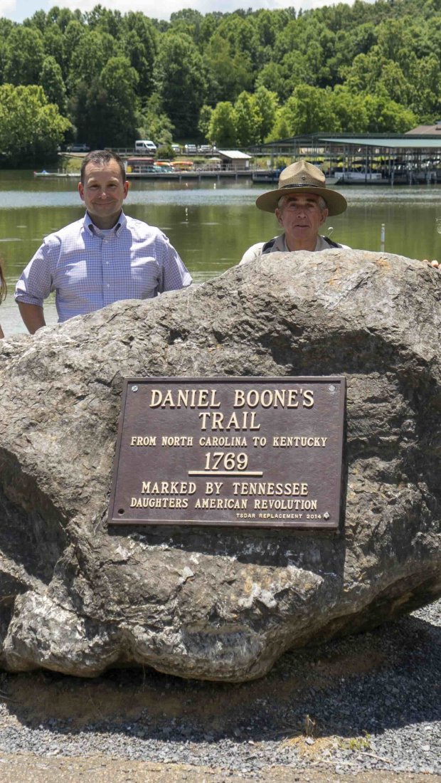 Four people stand by a large rock with a plaque marking "Daniel Boone's Trail" near a serene lake and lush green forest under a sunny sky.