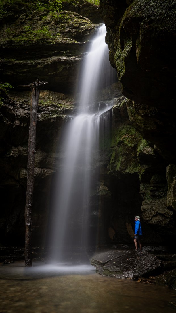 Waterfall cascading into a rocky cave with lush greenery, person in blue stands nearby.