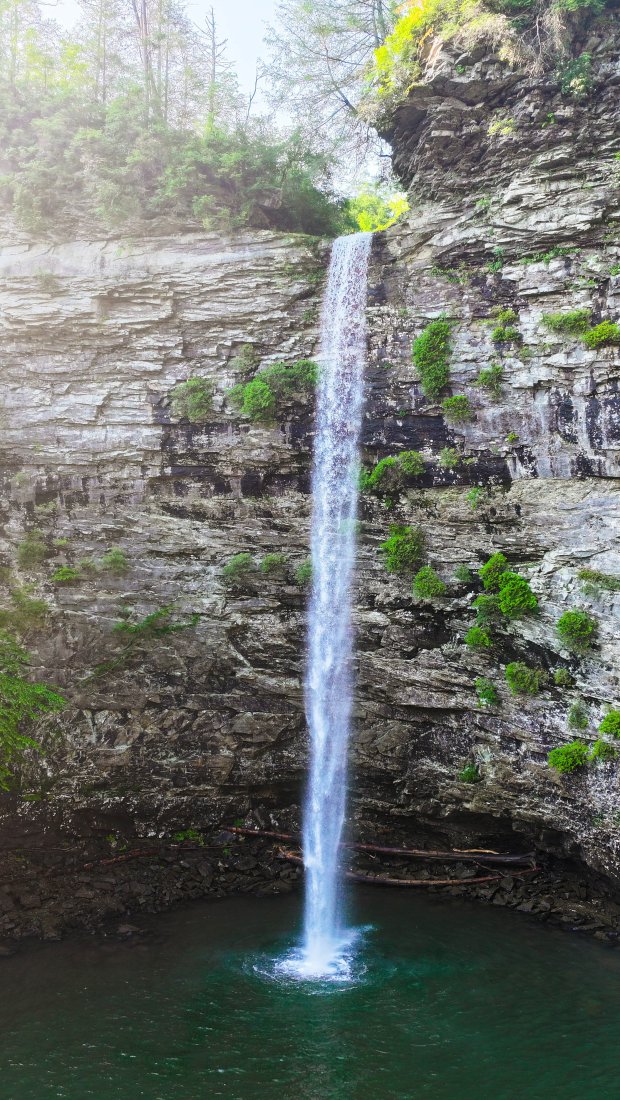 Tall waterfall cascading into a rocky pool surrounded by cliffs and trees.