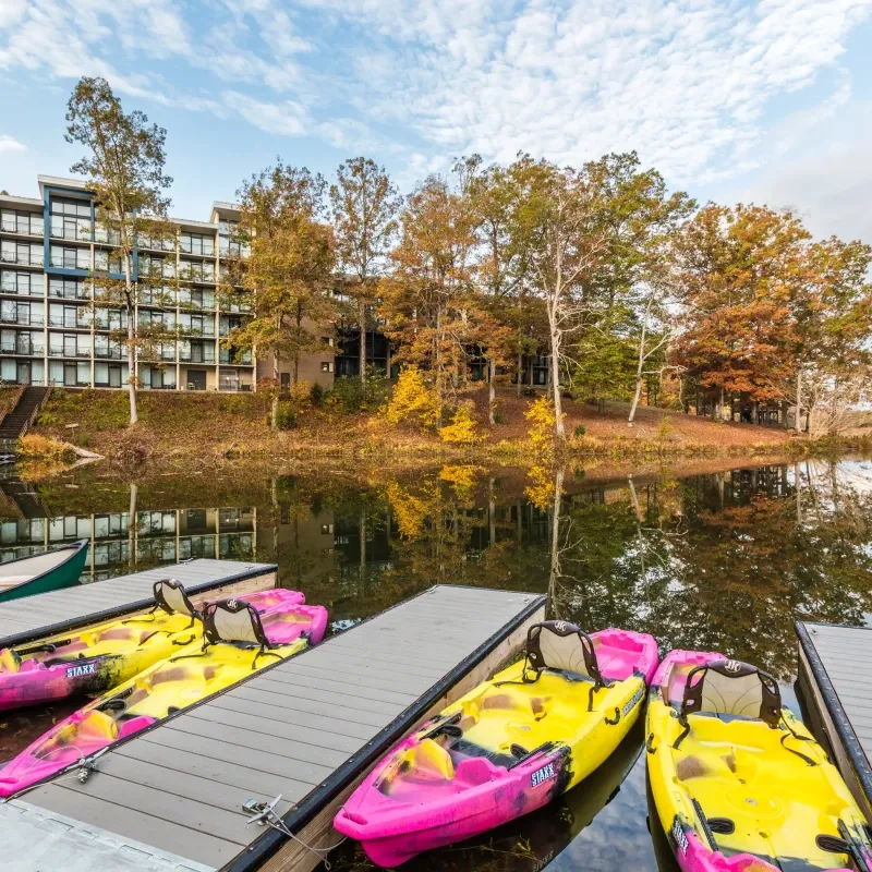 Kayaks docked by a calm lake, with a modern building and autumn trees in the background.