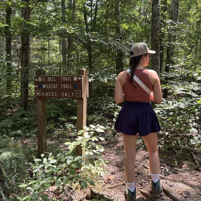 Hiker in woods near trail sign, wearing a cap and boots.