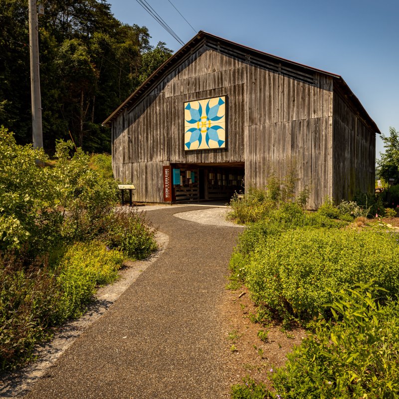 A rustic wooden barn with a colorful geometric quilt pattern on the front stands under a clear blue sky, surrounded by lush greenery and a winding path.