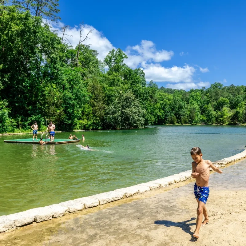 Boy in swim trunks on sandy shore, others swimming in a lake with lush green trees.