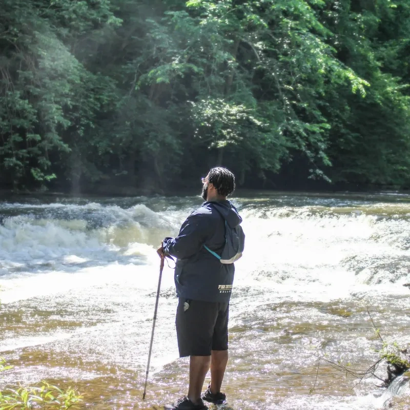 Person with hiking poles gazes at river, surrounded by lush green forest.