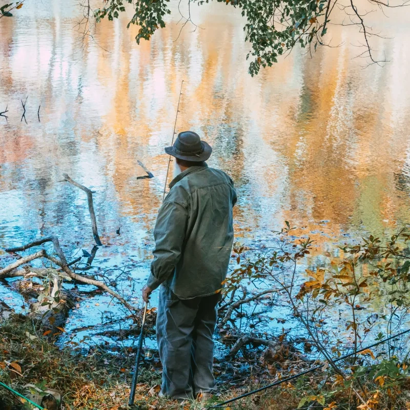 Man fishing by a calm, reflective lake surrounded by autumn trees.