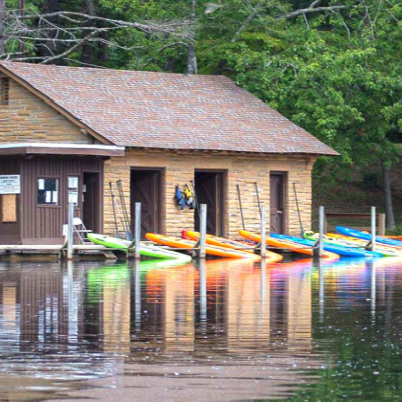A row of bright kayaks in red, orange, yellow, green, and blue line the dock outside a rustic stone boathouse on a calm lake.