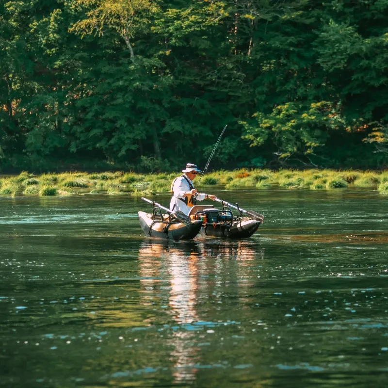 Person fishing from inflatable boat on a calm river, surrounded by lush trees.