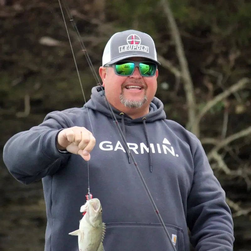 Man in sunglasses holding a caught fish, wearing a hoodie and cap, smiling outdoors.