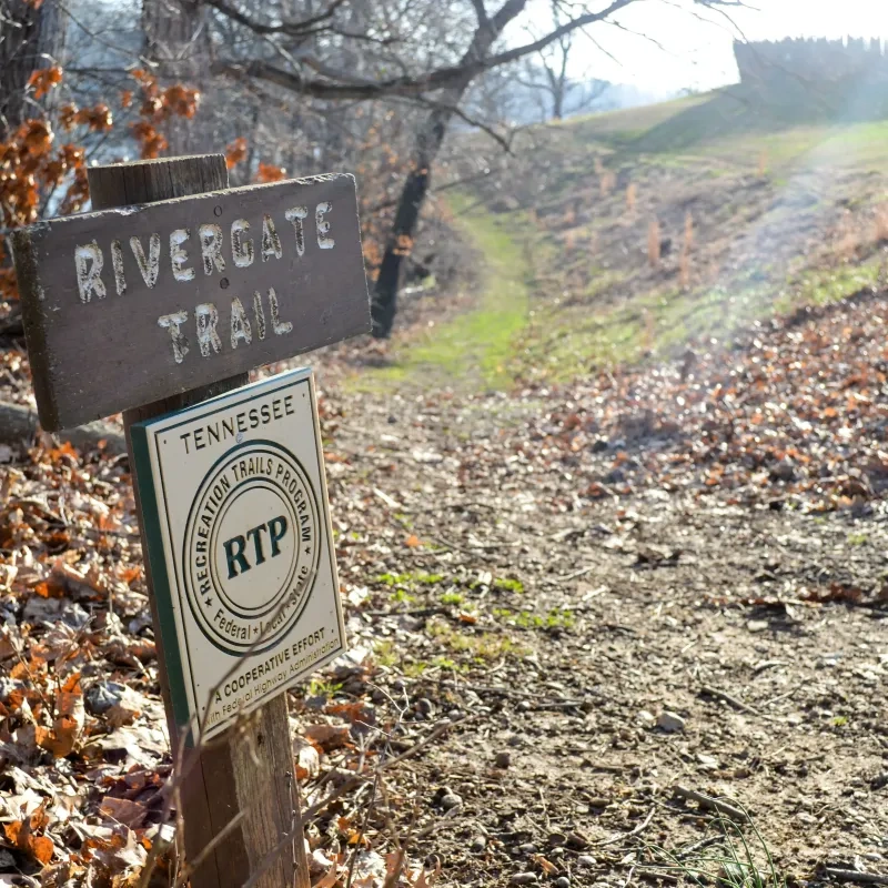 Trail sign in autumn leaves marks the Rivergate Trail in Tennessee. Sunlit path leads uphill, surrounded by bare trees and grassy patches.