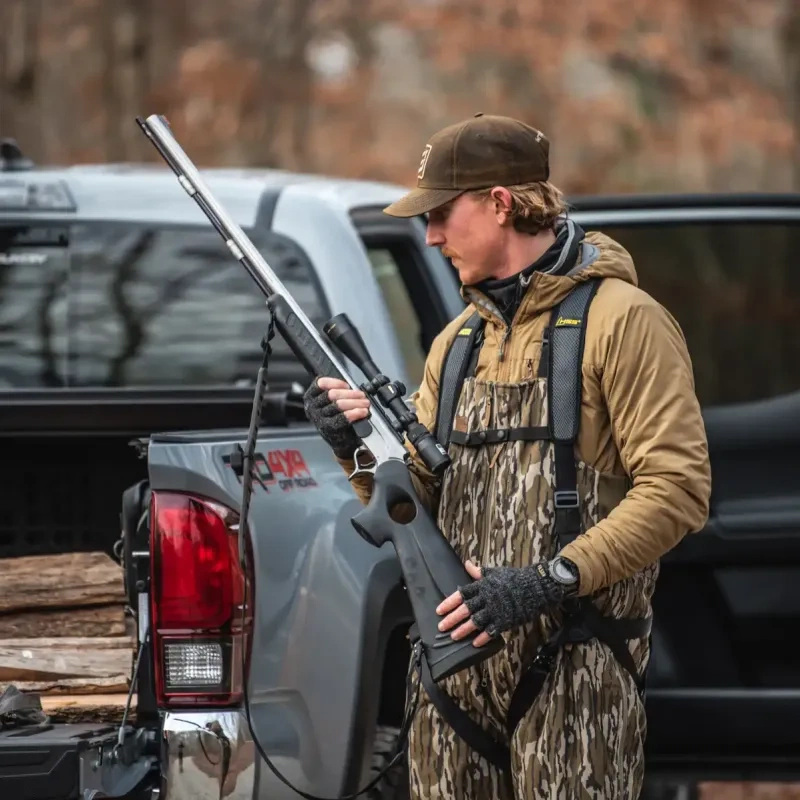 Hunter holding a rifle near a pickup truck in a wooded area.
