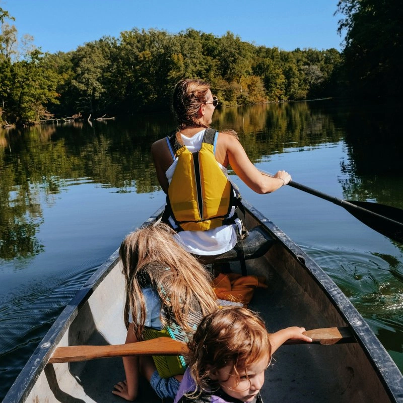 A woman and two children wearing life jackets paddle in a canoe on a calm river surrounded by lush green trees under a clear blue sky. Leisurely and serene.