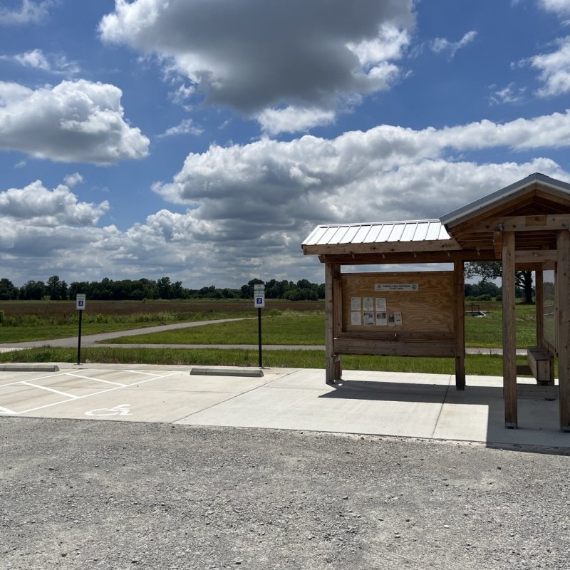 Wooden information kiosk under partly cloudy sky next to accessible parking lot, surrounded by green fields