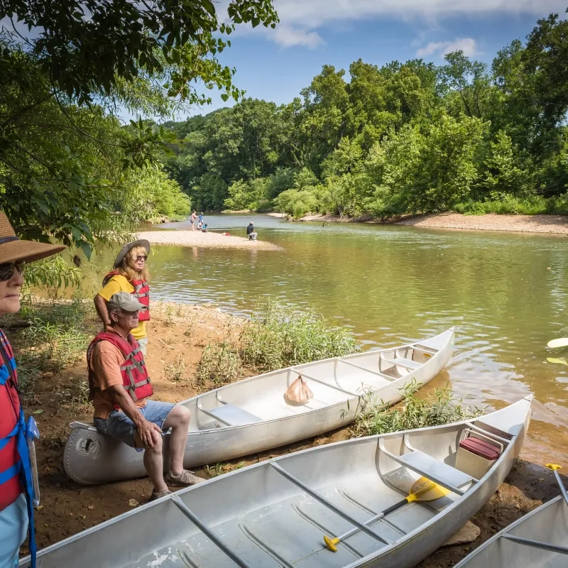 Canoeists in life vests by a riverbank, surrounded by greenery, with canoes on the shore.