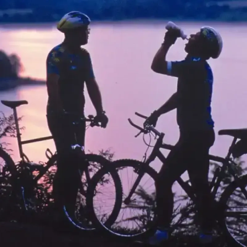 Two cyclists silhouette at sunset, one drinking water by a lake.