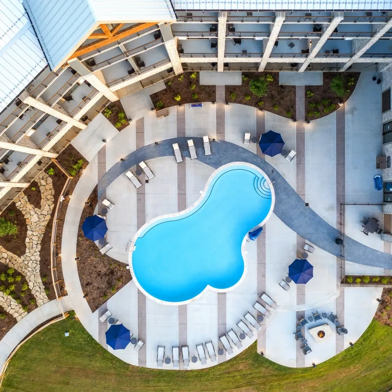 Aerial view of a curved pool with loungers and blue umbrellas on a spacious patio.
