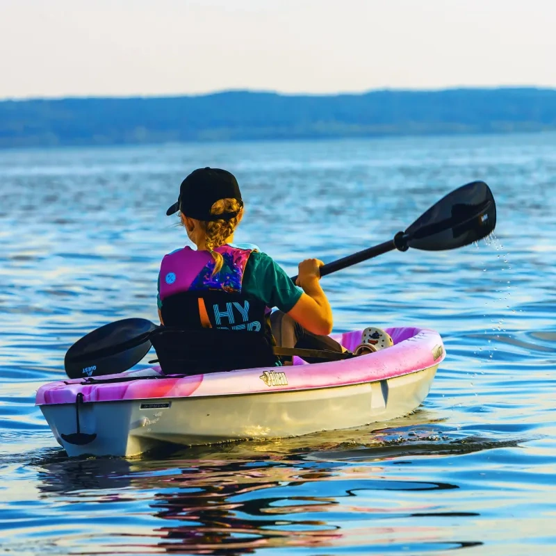 Person kayaking on a calm lake, wearing a cap and pink vest.