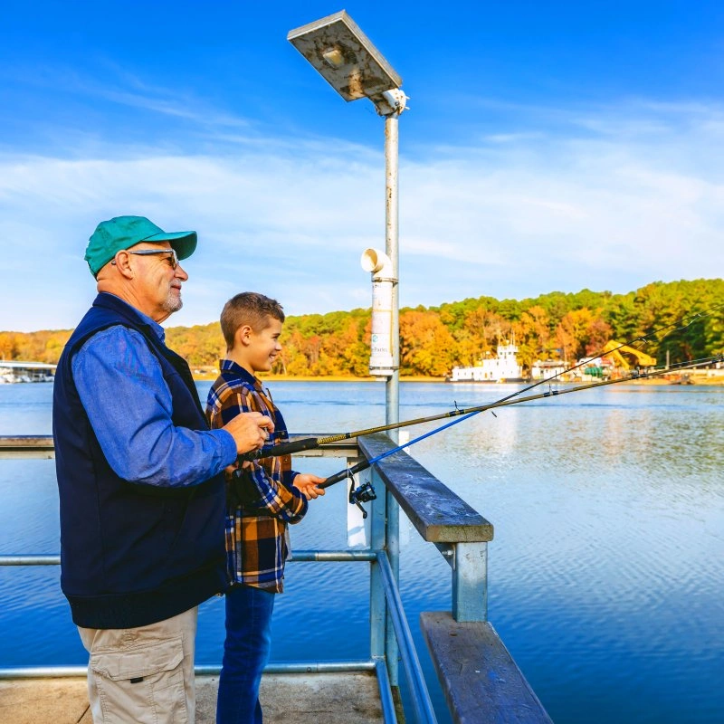 Man and boy fishing on a lakeside pier, surrounded by autumn trees.