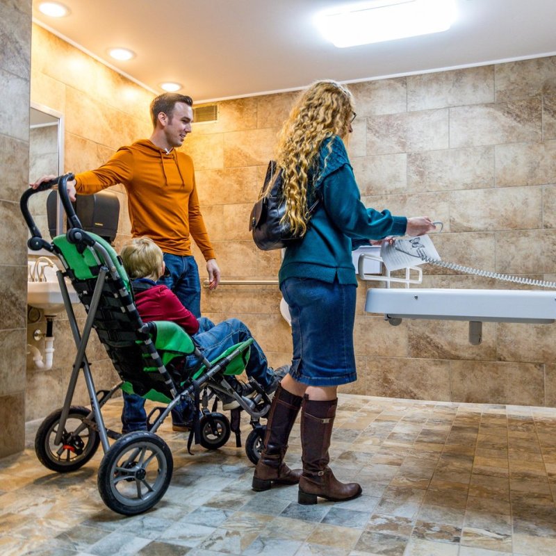 A family in a tiled bathroom with a changing table. A man holds a stroller with a child, while a woman interacts with the table. The scene is caring and relaxed.