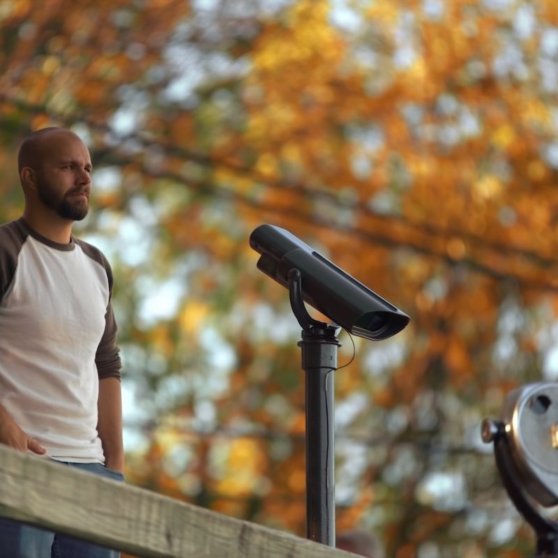 A man stands on a wooden deck, looking thoughtful beside a sightseeing telescope. Autumn trees with orange leaves create a warm, serene backdrop.