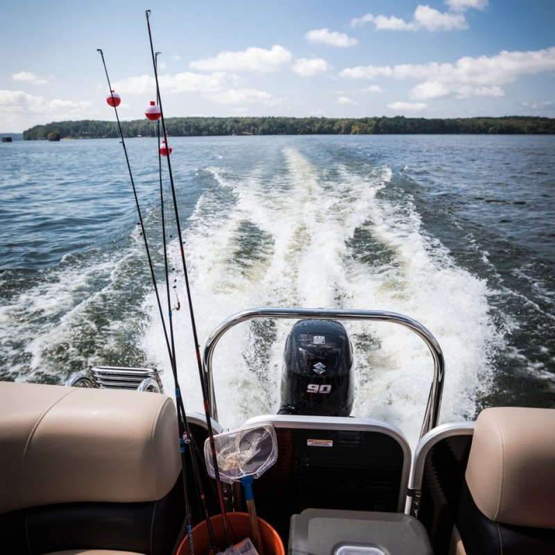 Boat on a lake under a bright blue sky, with fishing rods and gear onboard. Water churns behind, creating a lively and adventurous mood.