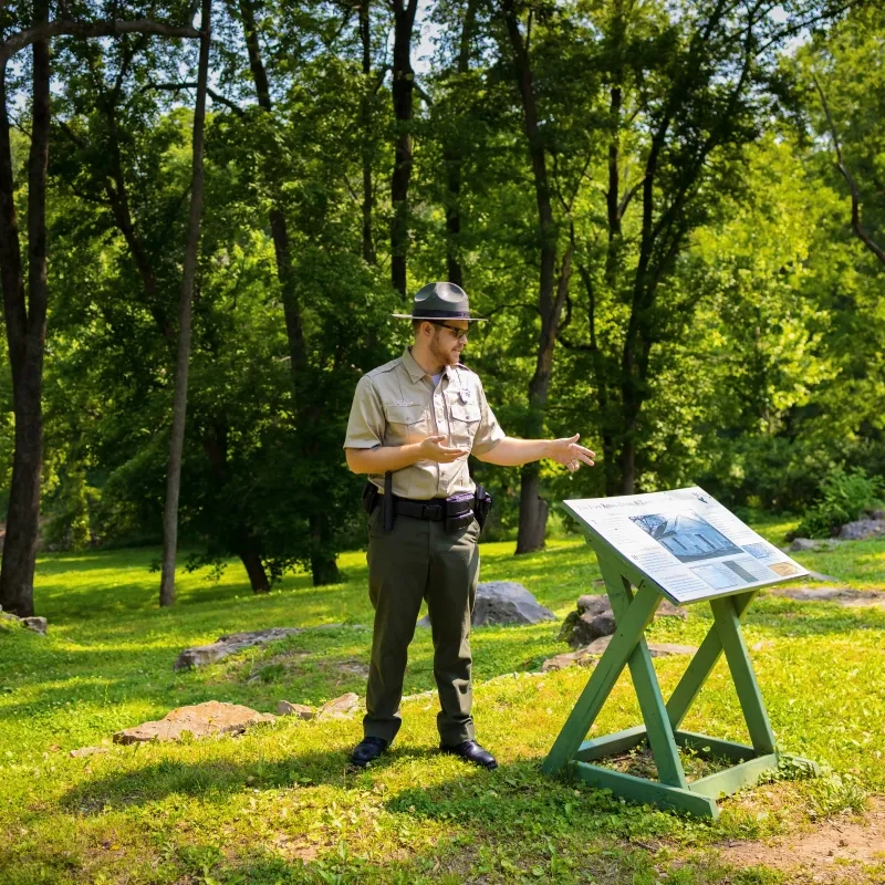 Park ranger gesturing at an informational sign in a lush, sunlit park.