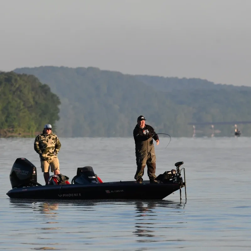 Two men fishing on a boat in a calm lake, surrounded by forested hills.